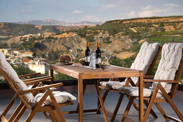 Mesa de madera con dos botellas de vino y copas, rodeada de sillas acolchadas, con vistas a un paisaje montañoso.