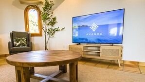 A room with a wooden table, a television displaying 'AVANOA', a chair with a leaf-patterned pillow, and a window with a plant. - A021 - COLINA DEL SOL (Calpe)