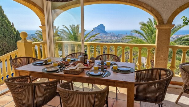 A dining table set with breakfast items, surrounded by wicker chairs, on a balcony with a view of a city and mountains. - A021 - COLINA DEL SOL (Calpe)