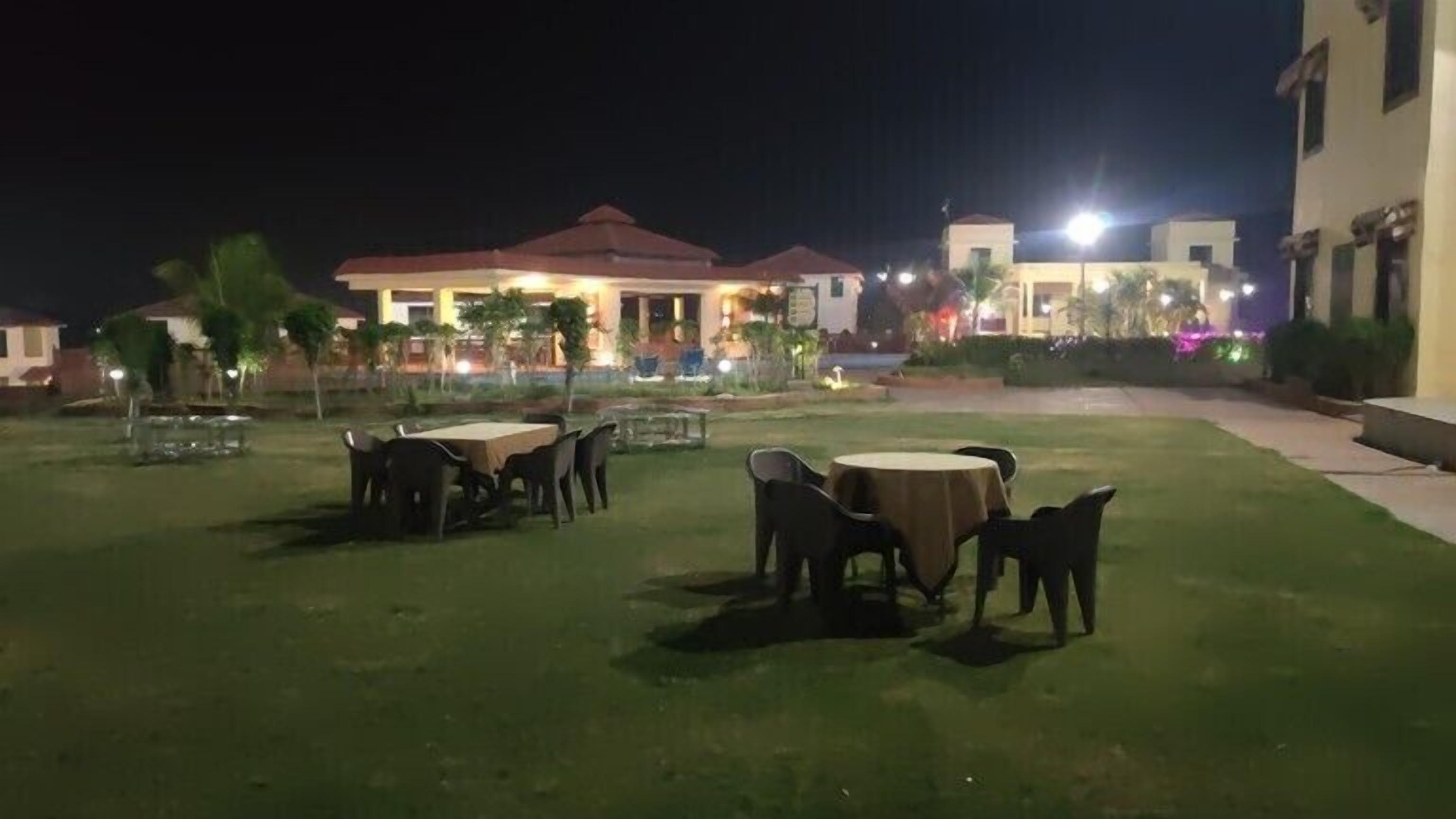 An outdoor seating area with tables and chairs, a building with a red roof in the background, and a well-lit pathway.