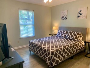 A bedroom with a bed, a TV, a window with blinds, and two wall art pieces.