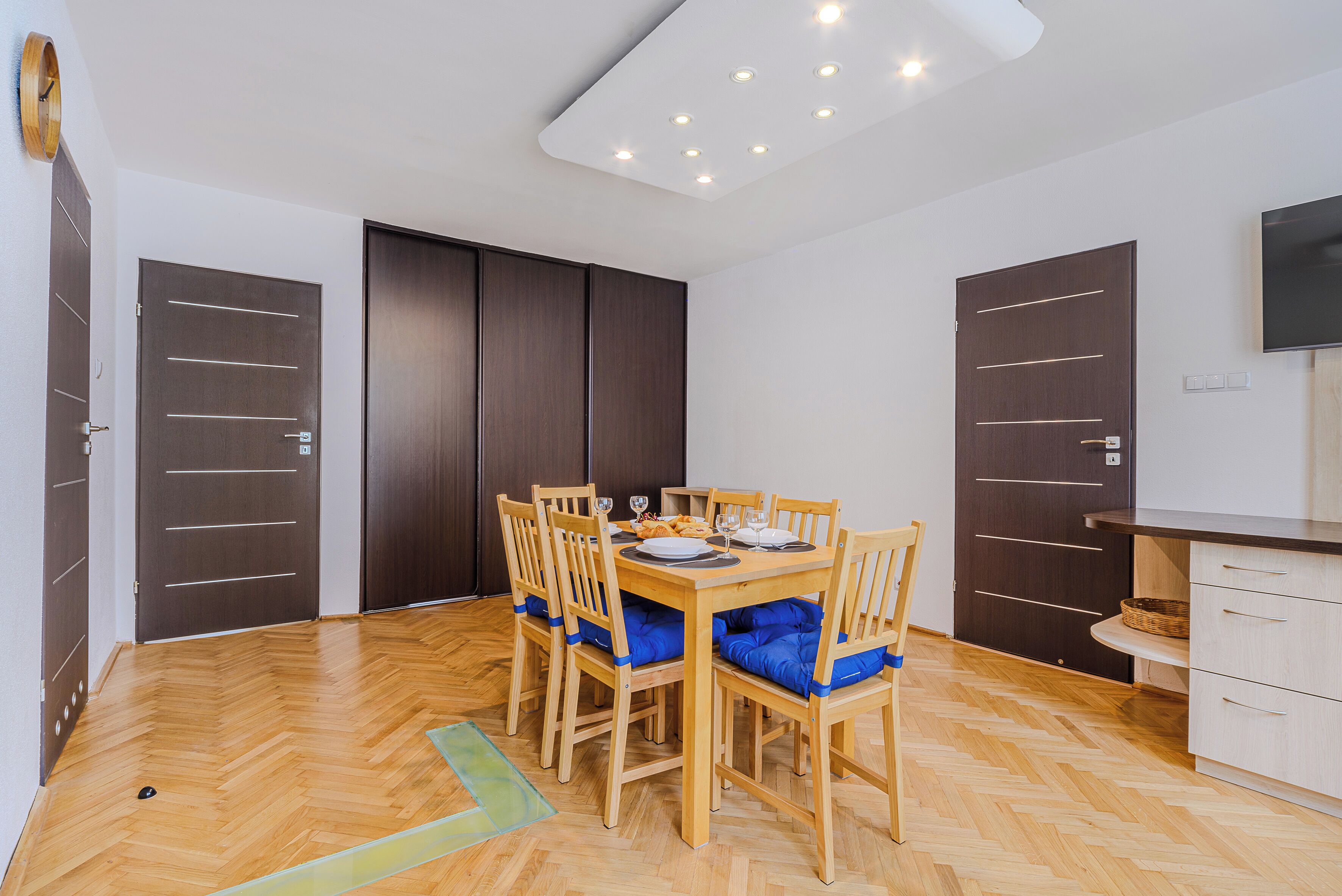 Un comedor con mesa y sillas de madera, un televisor en la pared y una zona de cocina con armarios y encimera.