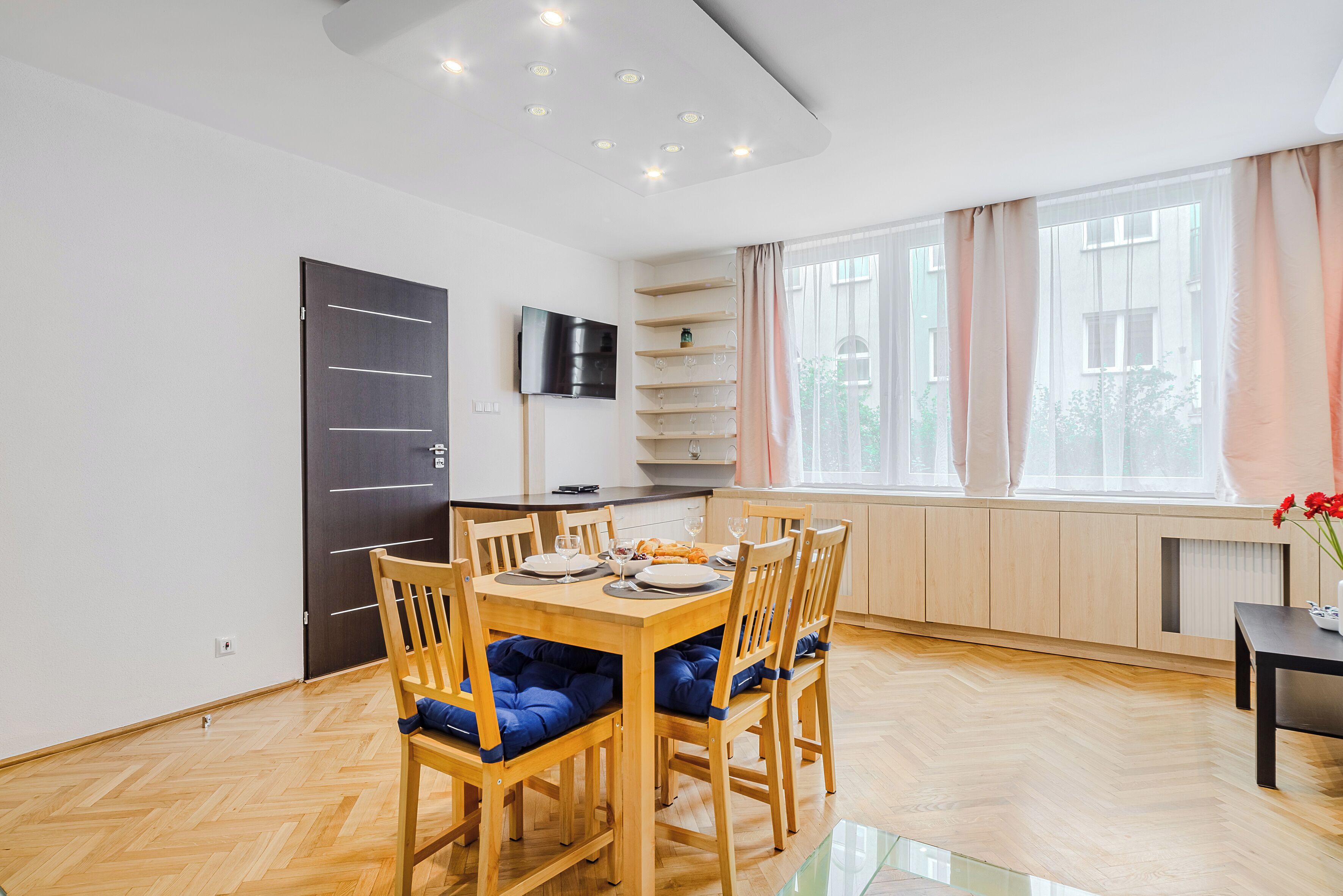 Un coin repas avec une table et des chaises en bois, un téléviseur fixé au mur et une fenêtre avec des rideaux.