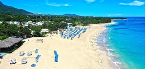 Una spiaggia con file di ombrelloni blu e lettini, un edificio con tetto di paglia e vista sull'oceano e sulle montagne sullo sfondo.