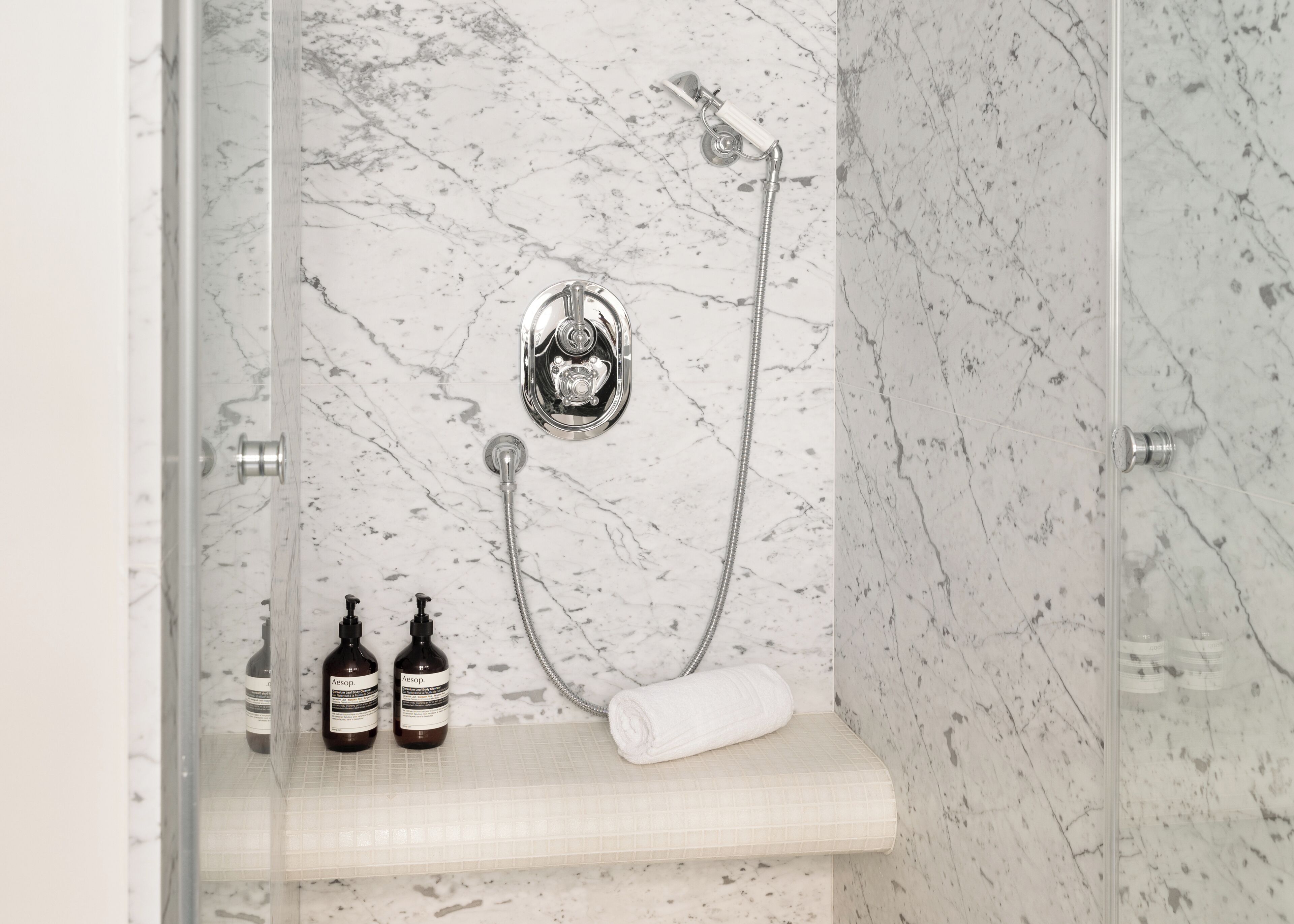 A bathroom with a glass-enclosed shower, marble walls, and a shelf with two bottles and a rolled towel.