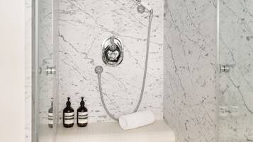A bathroom with a glass-enclosed shower, marble walls, and a shelf with two bottles and a rolled towel.