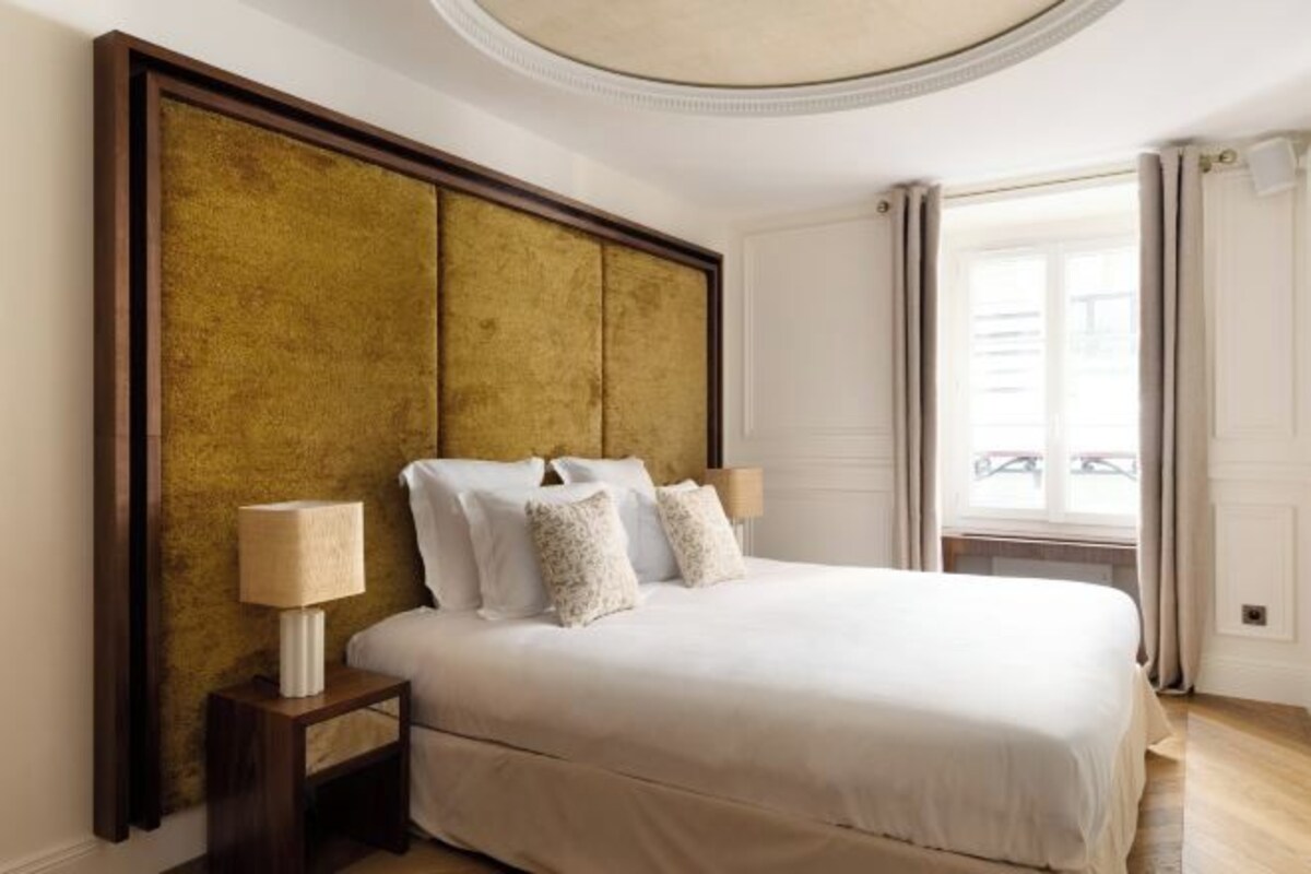 Une chambre à coucher avec un grand lit, deux tables de chevet avec des lampes, une tête de lit dorée et une fenêtre avec des rideaux.