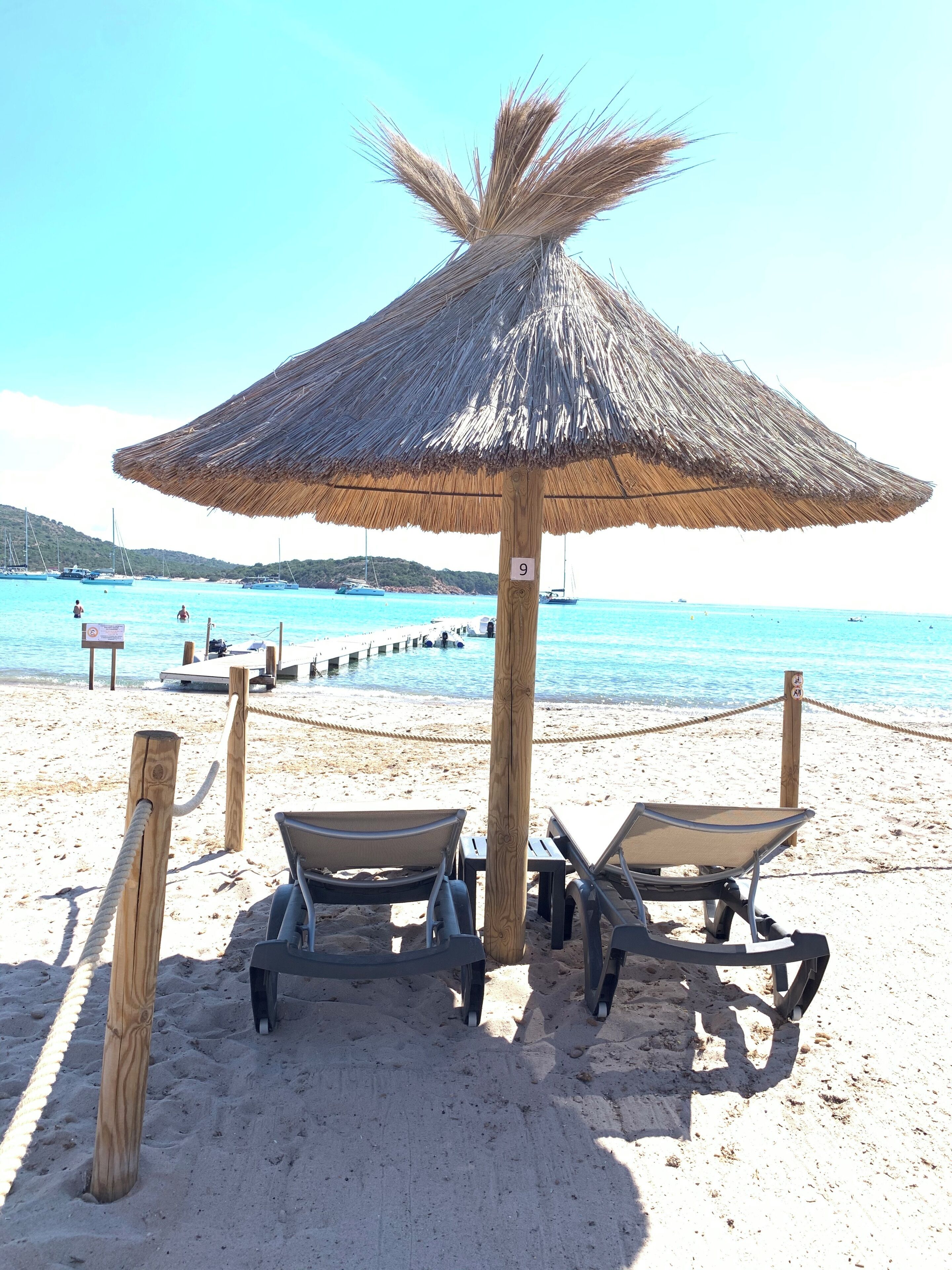 A beach with a thatched umbrella and two lounge chairs.