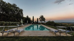 Et rektangulært swimmingpool med liggestole og parasoller på hver side, omgivet af græs og træer, med udsigt til fjerne bakker.