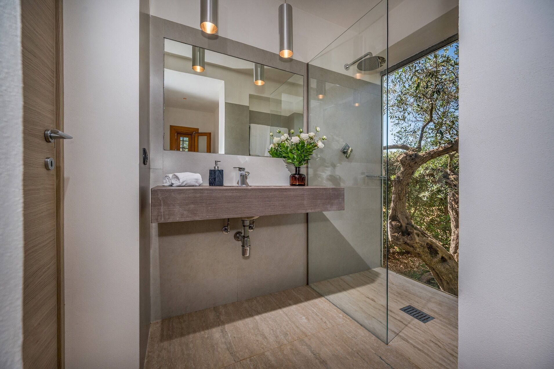 Un bagno moderno con box doccia in vetro, mobile lavabo in legno, uno specchio grande e vista su alberi.