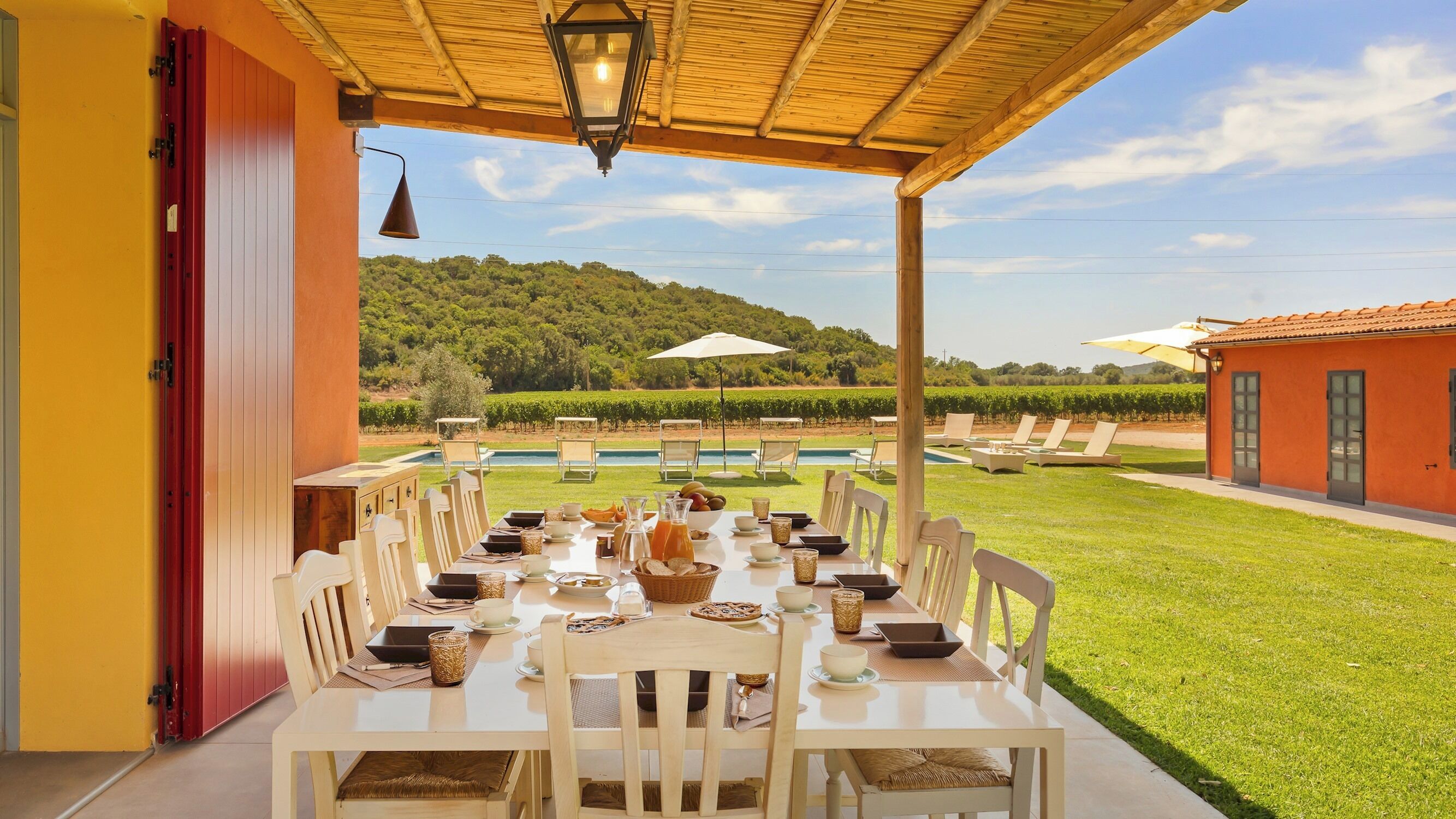 A covered outdoor dining area with a table set for a meal, overlooking a vineyard and a swimming pool.