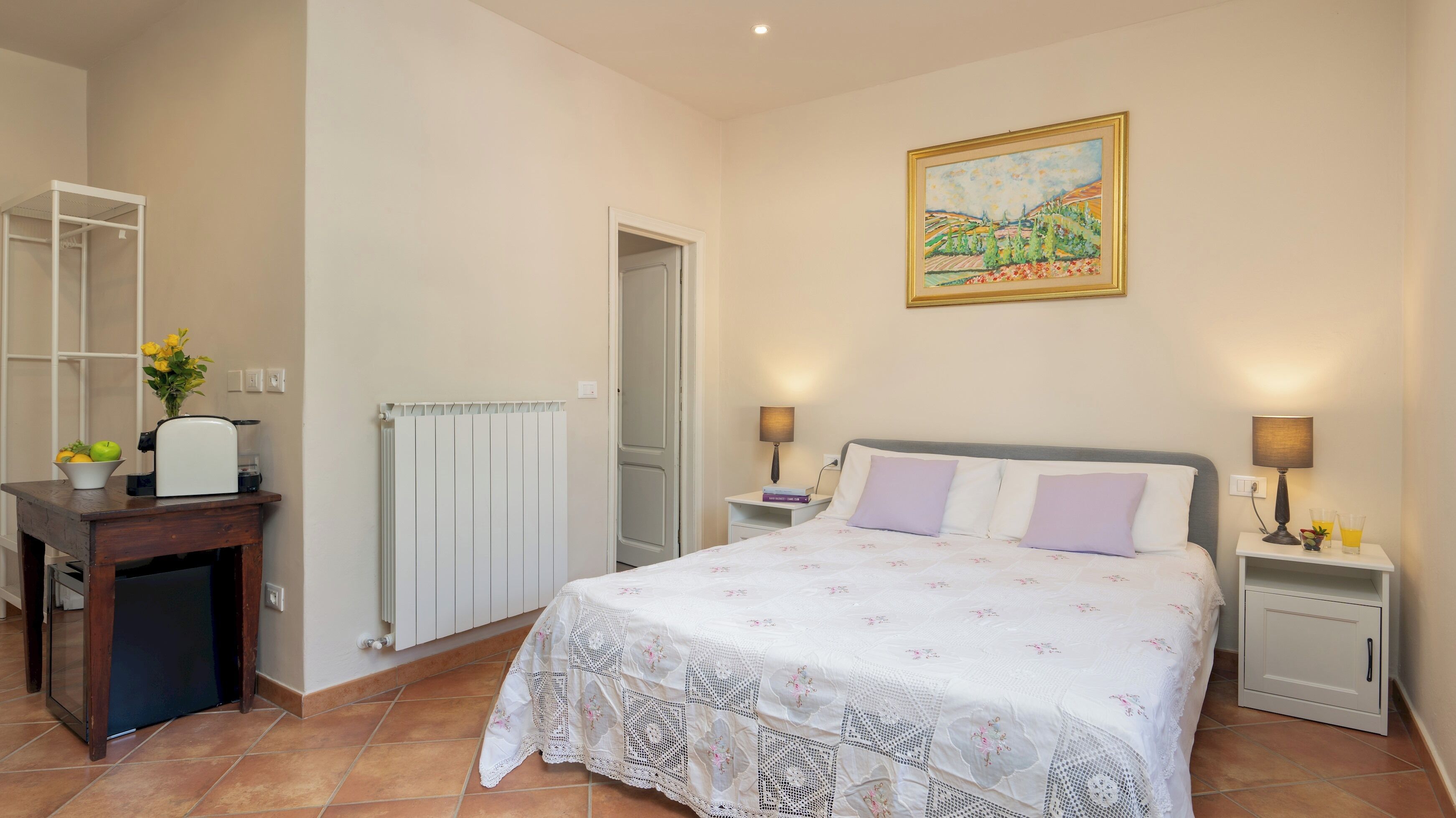 A bedroom with a bed, bedside tables, a radiator, a desk with a coffee maker, and a painting on the wall.