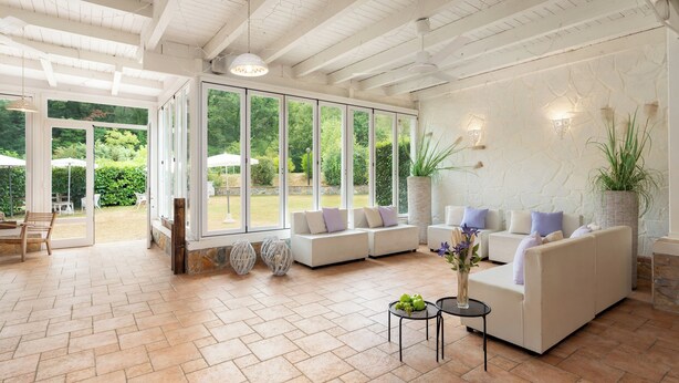 Uma sala de estar espaçosa com uma grande porta de vidro que dá para uma área externa, sofás brancos com almofadas, uma pequena mesa redonda com um vaso e uma planta em vaso.