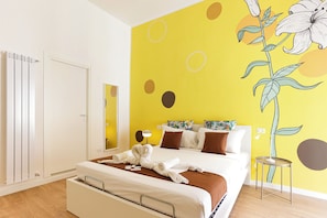 Une chambre d’hôtel comprenant un lit, une table de chevet, un miroir, un radiateur et un mur orné d’une fresque florale.