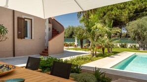 Zona junto a la piscina con mesa de madera, sillas y una sombrilla grande. Se ve una escalera que sube a un nivel superior, rodeado de vegetación y una piscina.