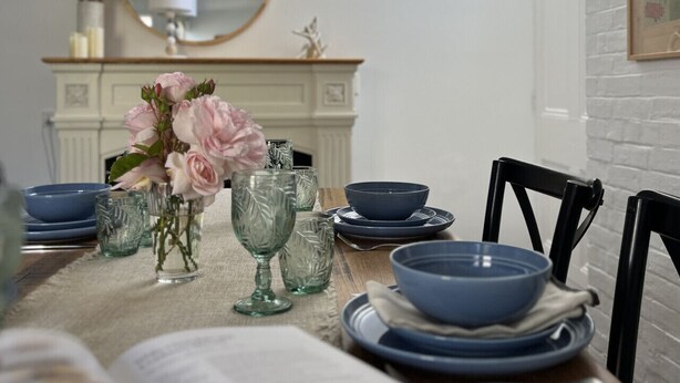 A dining table set with blue dishes and glassware, a vase with pink flowers, and a mirror above.