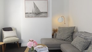 A living room with a grey sofa, a chair, a coffee table with books and flowers, a framed picture on the wall, and a lamp.