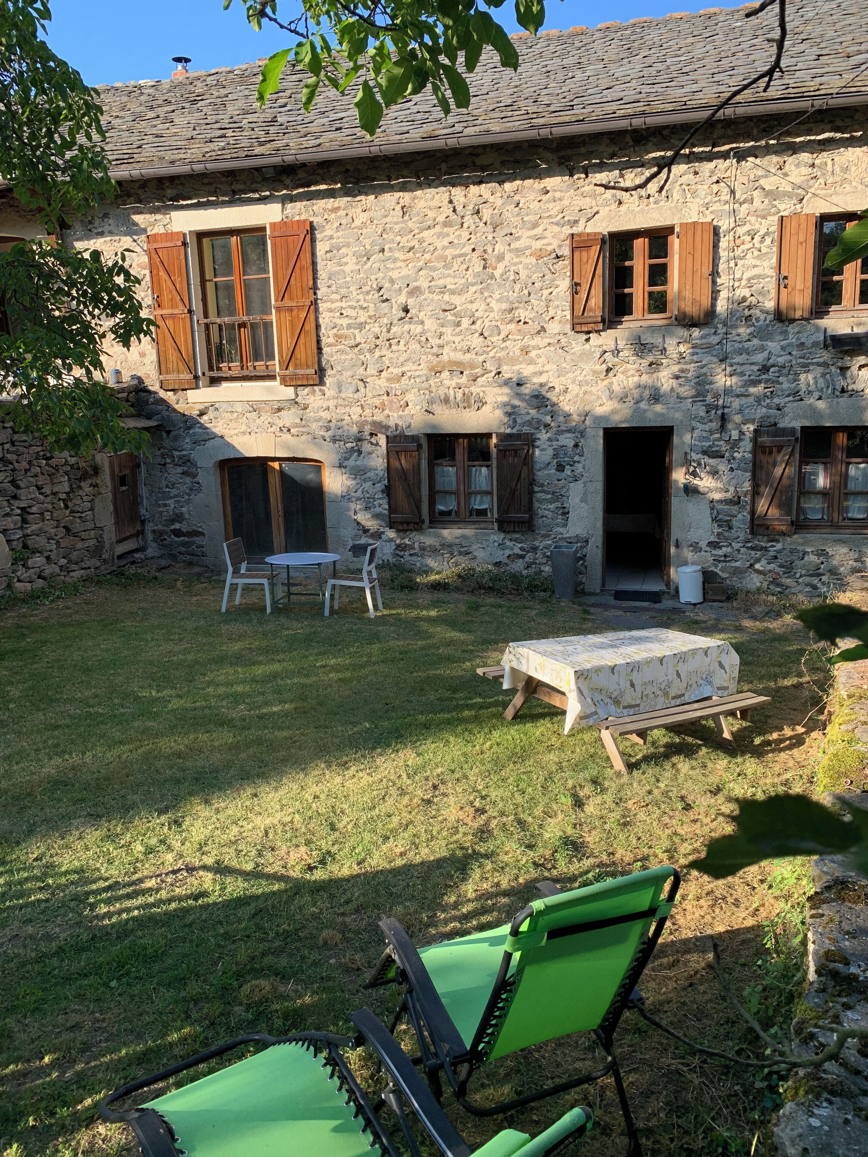 Un bâtiment en pierre avec des volets en bois, un petit patio avec une table et des chaises, et une pelouse verdoyante.