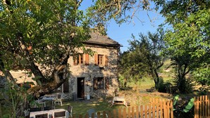 Un bâtiment en pierre avec une clôture en bois, des sièges à l’extérieur et un arbre au premier plan.