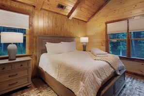 Un dormitorio de cabaña de madera con una cama, mesitas de noche, lámparas y una ventana con vistas a árboles.