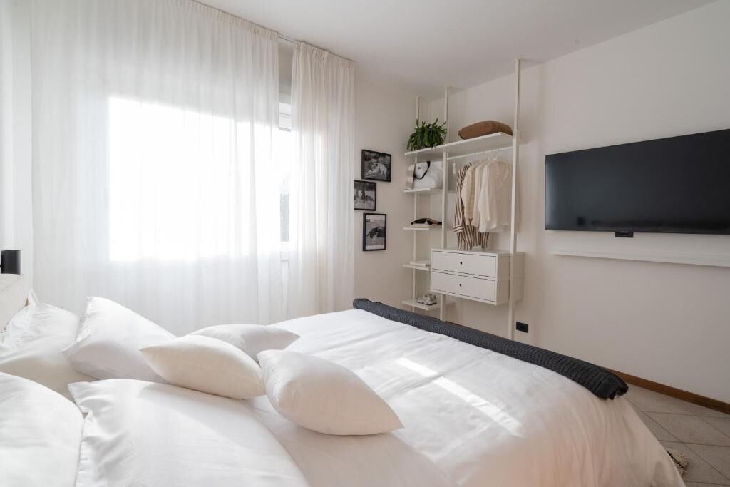 A bedroom with a large bed, white bedding, a TV mounted on the wall, a wardrobe, and a window with sheer curtains.