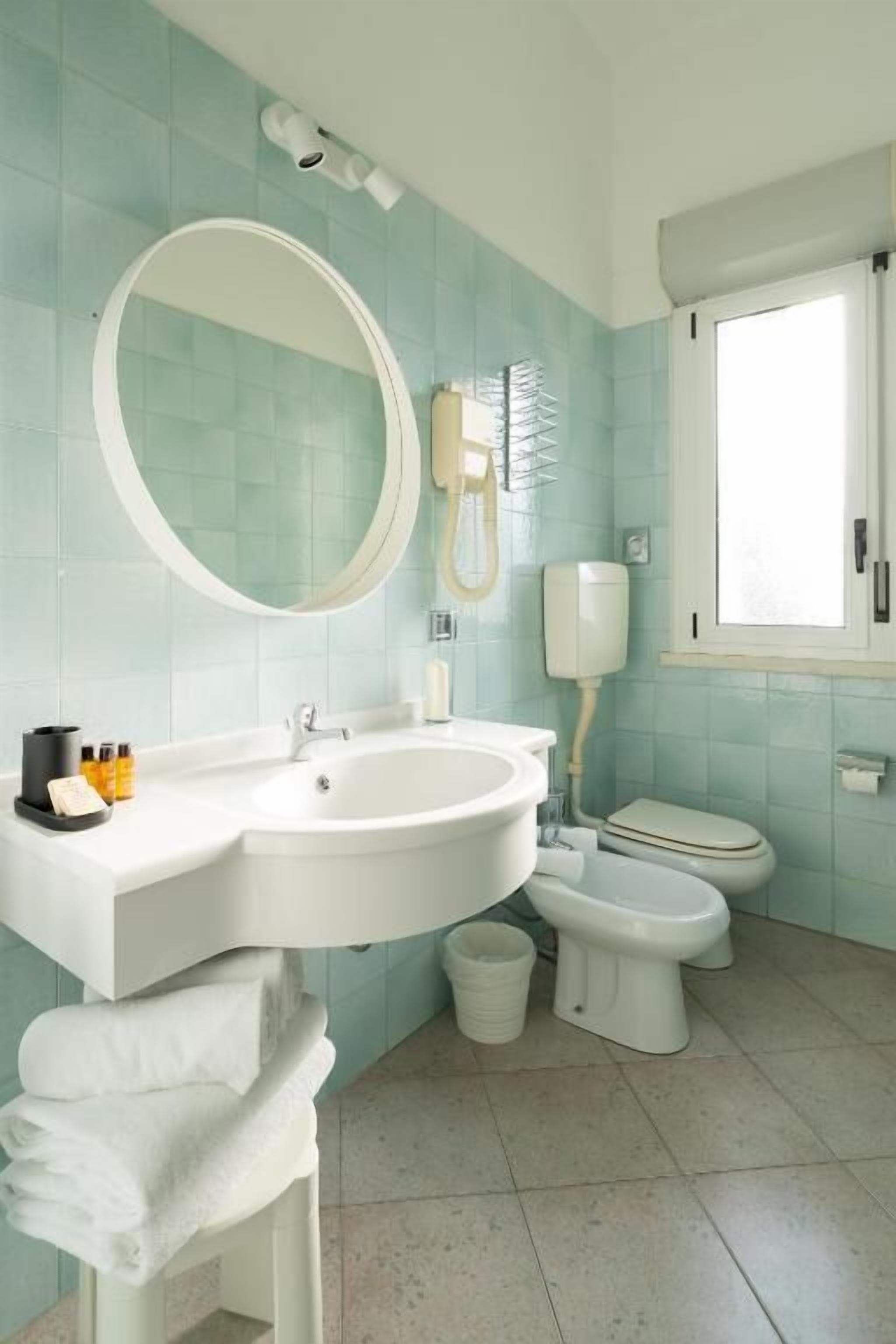 Un baño con lavamanos, espejo y retrete blancos. Hay una ventana que proporciona luz natural.
