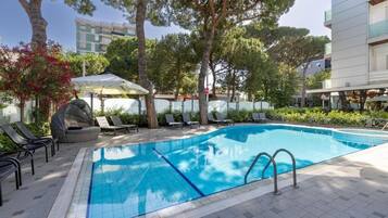 Une piscine entourée d’arbres et d’un bâtiment, avec des chaises longues et des parasols.