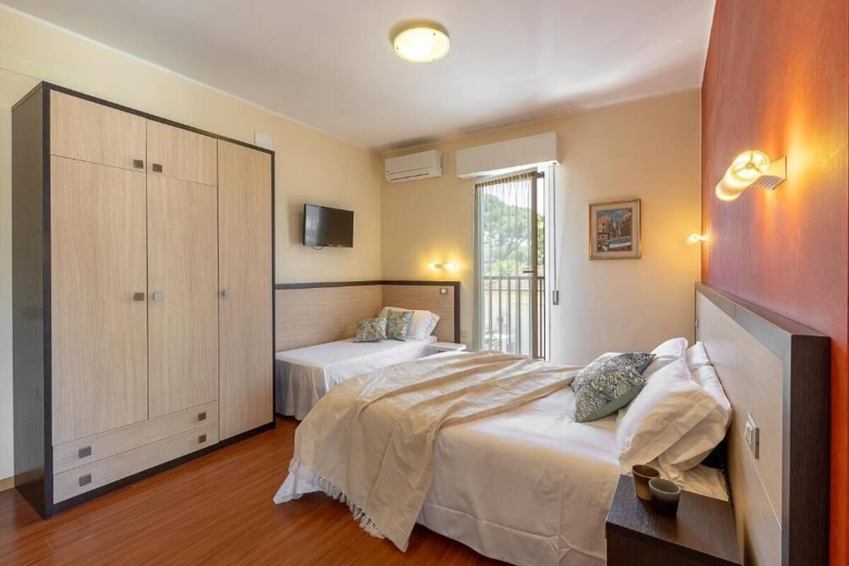 Een slaapkamer met twee bedden, een kledingkast, een televisie en een raam met jaloezieën.