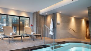 A modern spa area with a glass-enclosed hot tub, two lounge chairs, and a view of the city through large windows.