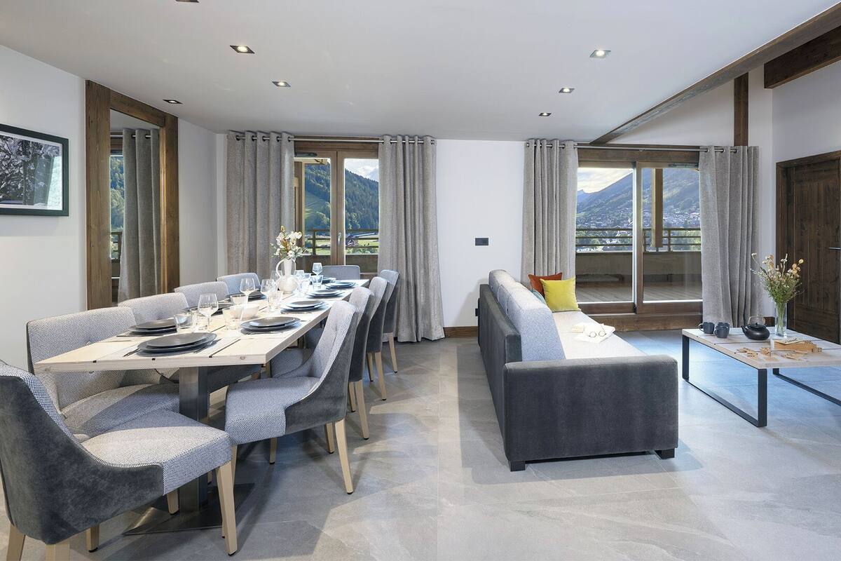 A modern hotel room with a dining area, a sofa, a TV, and a balcony with a mountain view.