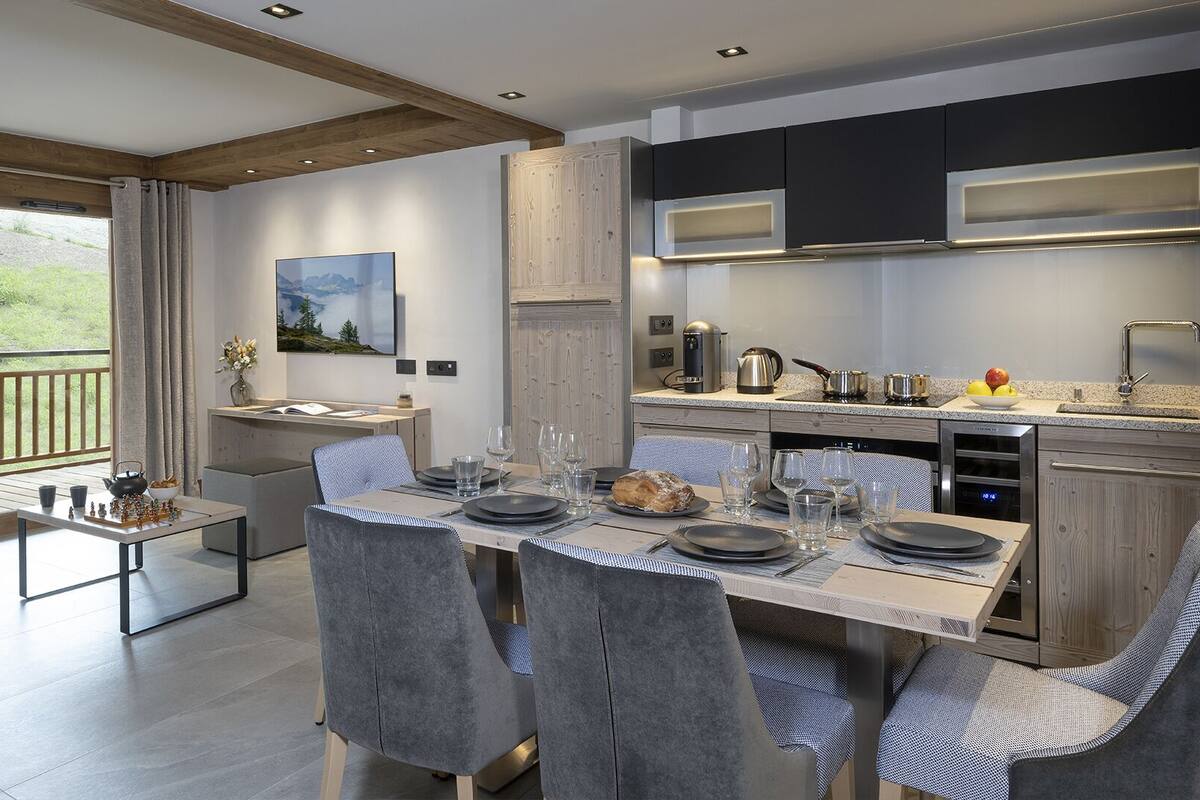 A modern kitchen with integrated appliances, a dining area with a table set for four, and a balcony with a view of greenery.