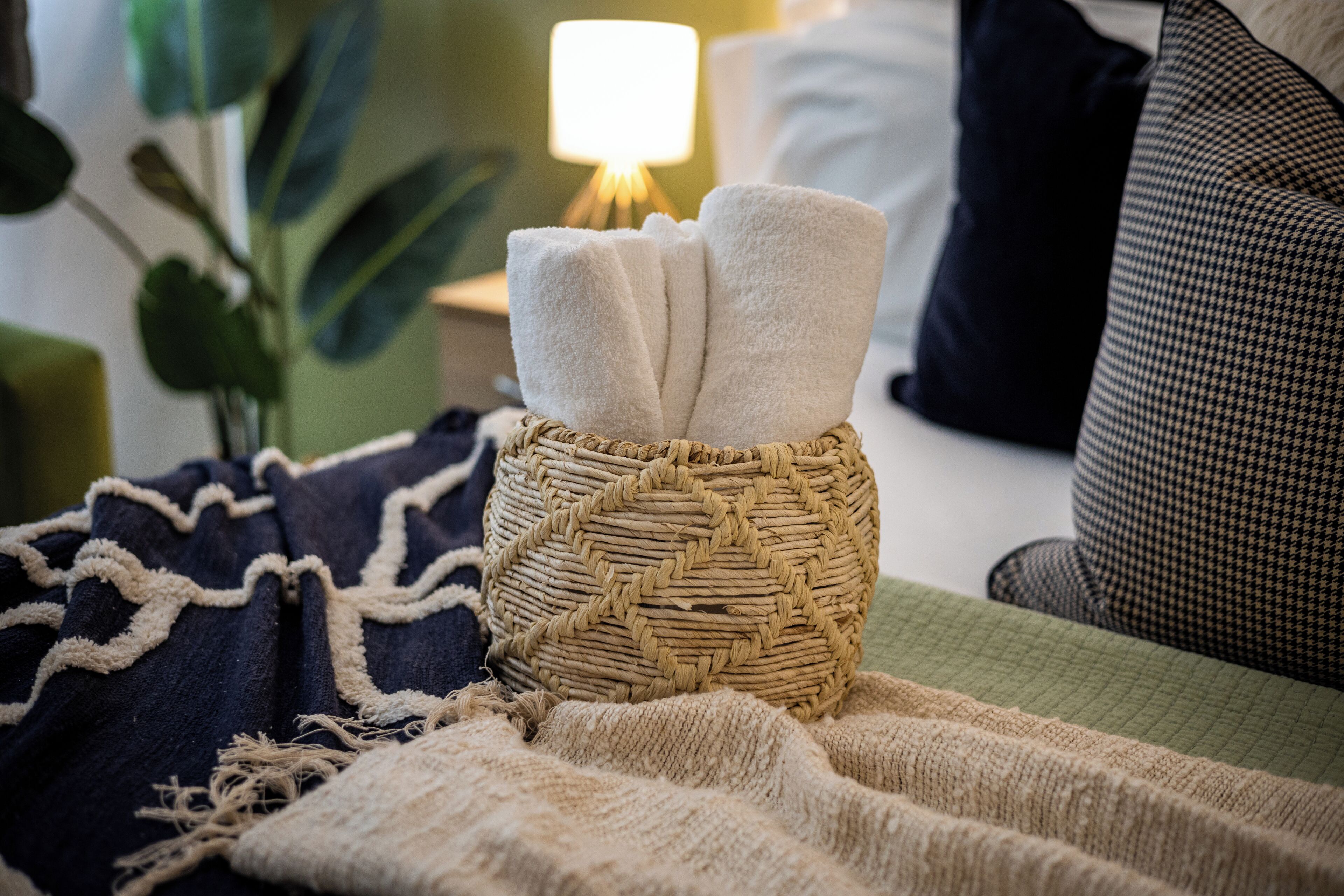A cozy room with a woven basket holding towels, a green blanket, and a bed with pillows.