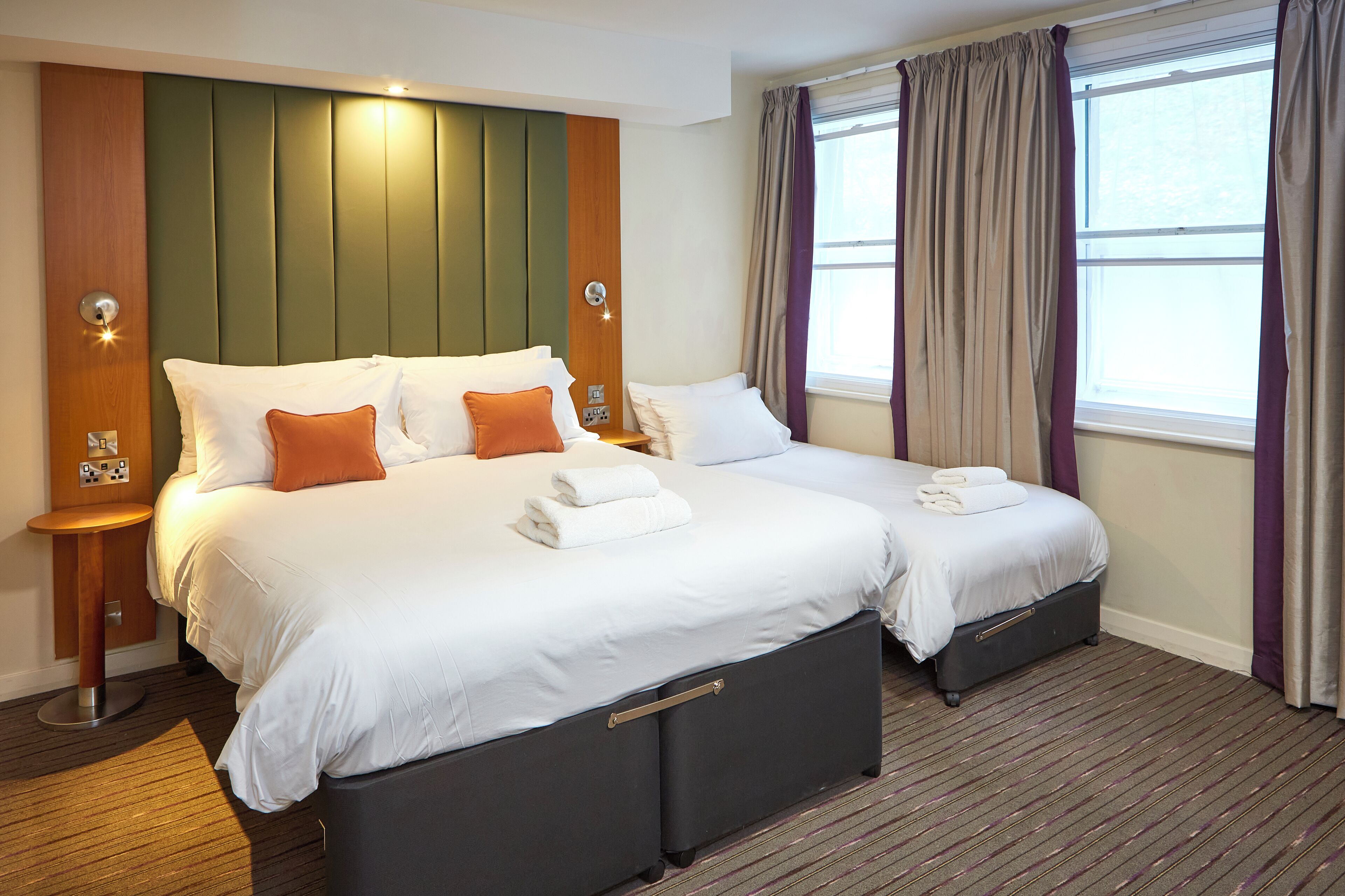 A hotel room with two beds, a wooden headboard, a small table, and a window with curtains.