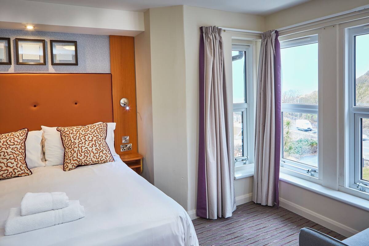 Une chambre d’hôtel avec un grand lit, une tête de lit, une table de chevet, une fenêtre avec des rideaux et une vue sur l’extérieur.