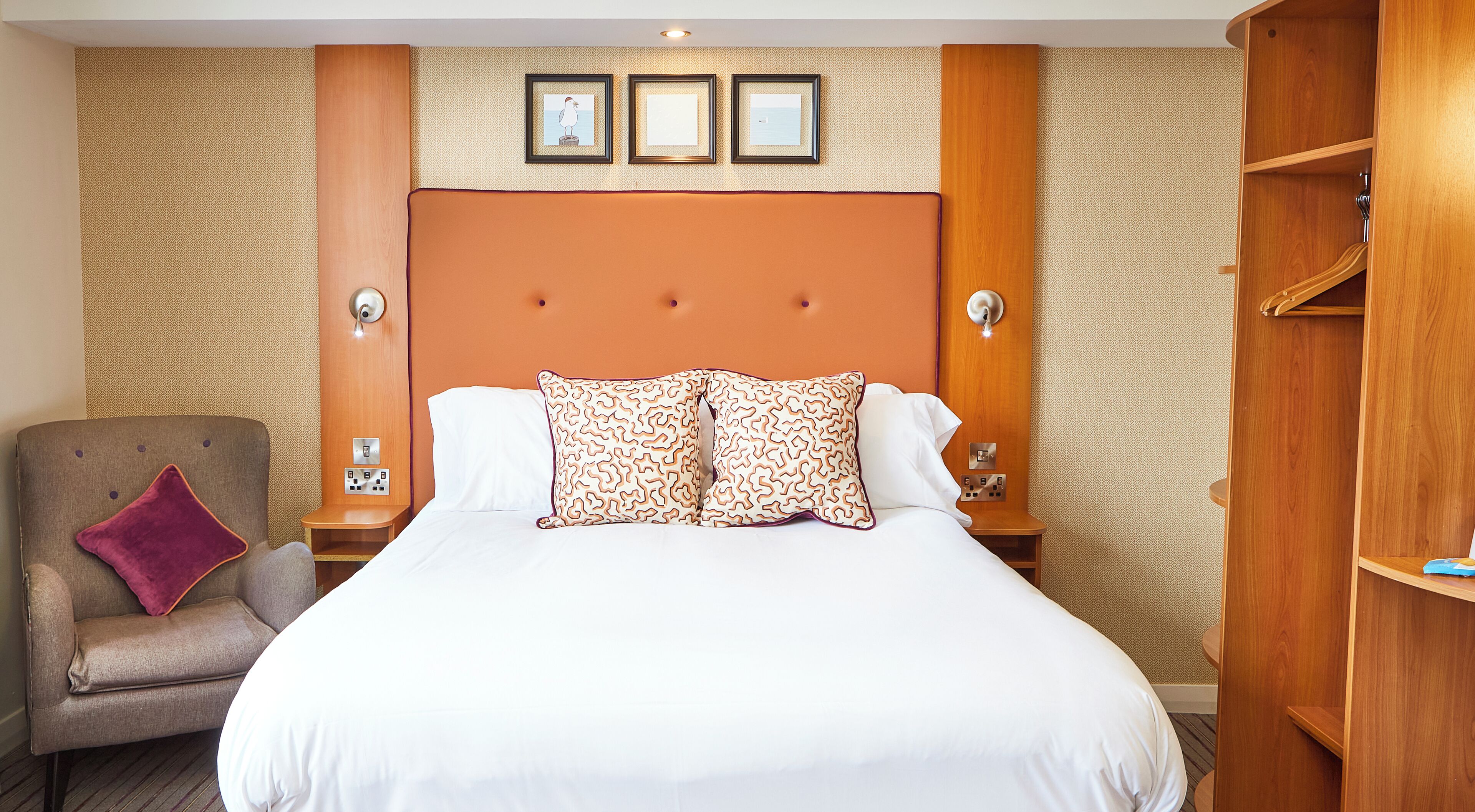 Une chambre d’hôtel avec un grand lit, une tête de lit en bois, une table de chevet avec une lampe, une chaise avec un coussin et une armoire.