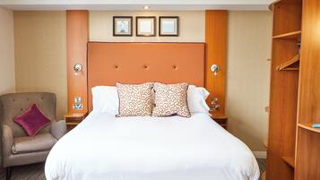 Habitación de hotel con una cama grande, un cabecero de madera, una mesita de noche con lámpara, un sillón con cojín y un armario.