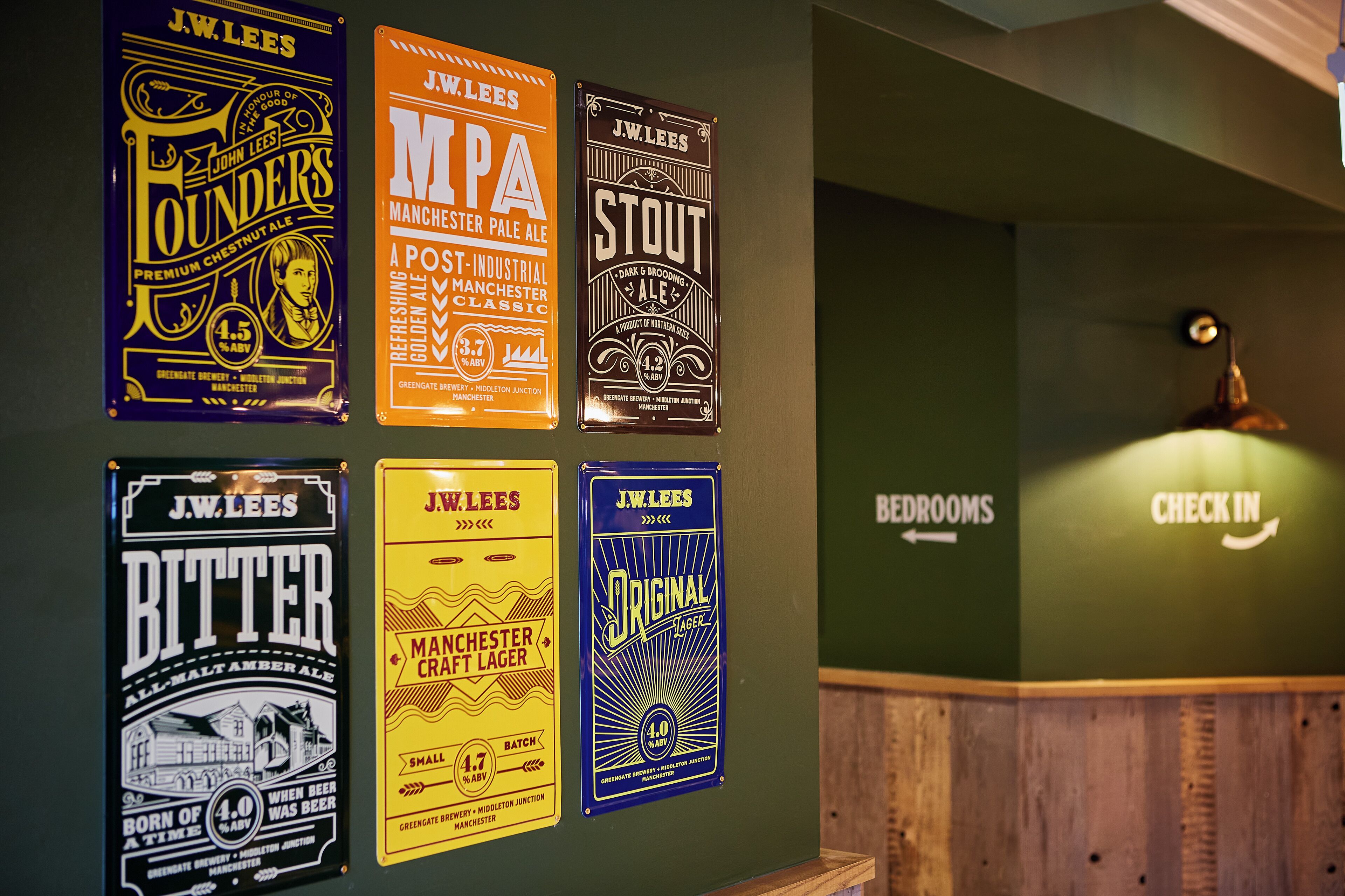 Eine Wand mit verschiedenen J.W. Lees Bierplakaten und einem Schild mit den Hinweisen 'ZIMMER' und 'CHECK IN'.