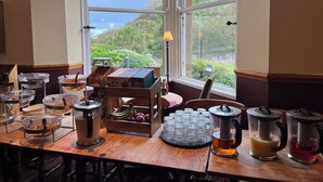 A breakfast buffet setup with various food items and beverages, a view of a hillside through large windows. - Craigside Manor (Llandudno)