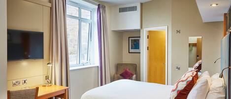 A hotel room with a bed, a desk with a lamp, a chair, a television, and a window with curtains.