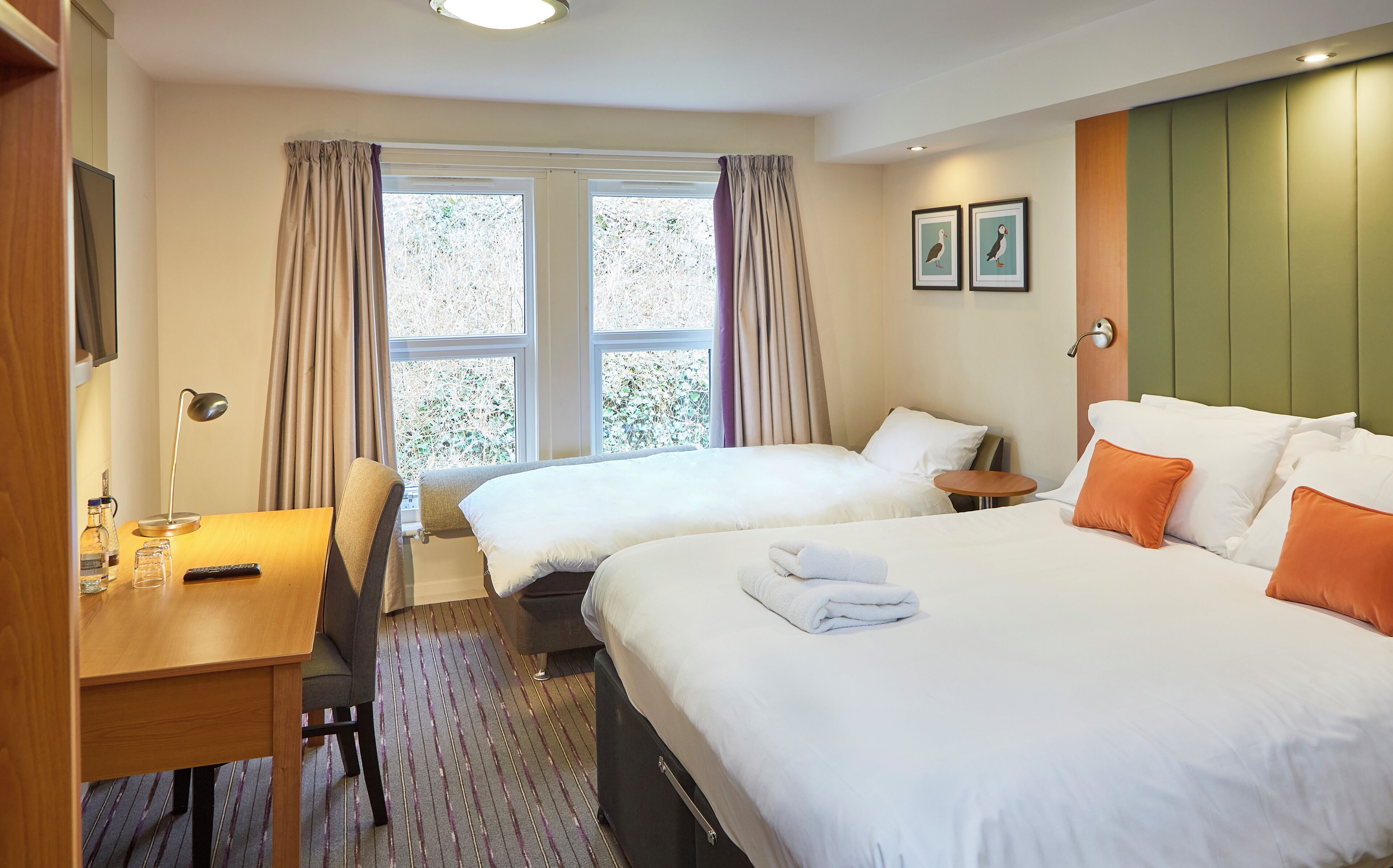 Une chambre d’hôtel avec deux lits, un bureau, une chaise, une petite table et une fenêtre avec des rideaux.