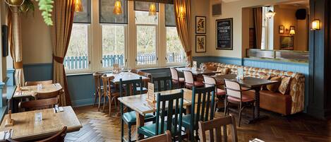 A restaurant interior with wooden tables and chairs, a blue patterned bench, and large windows with curtains.