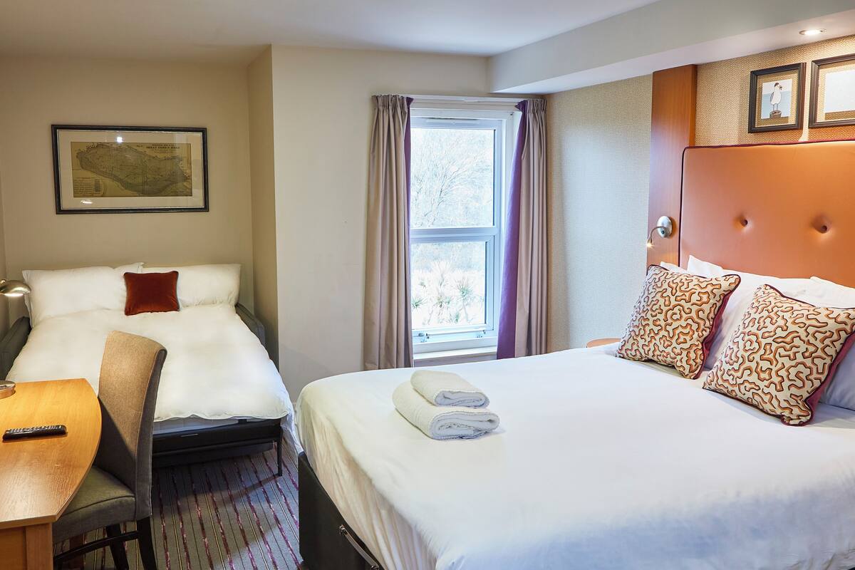 Une chambre d’hôtel avec deux lits, un bureau, une chaise, une fenêtre avec des rideaux et des tableaux encadrés au mur.