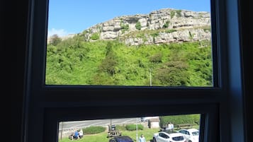 Vista de un estacionamiento con coches y gente, una zona con césped y una colina rocosa al fondo.
