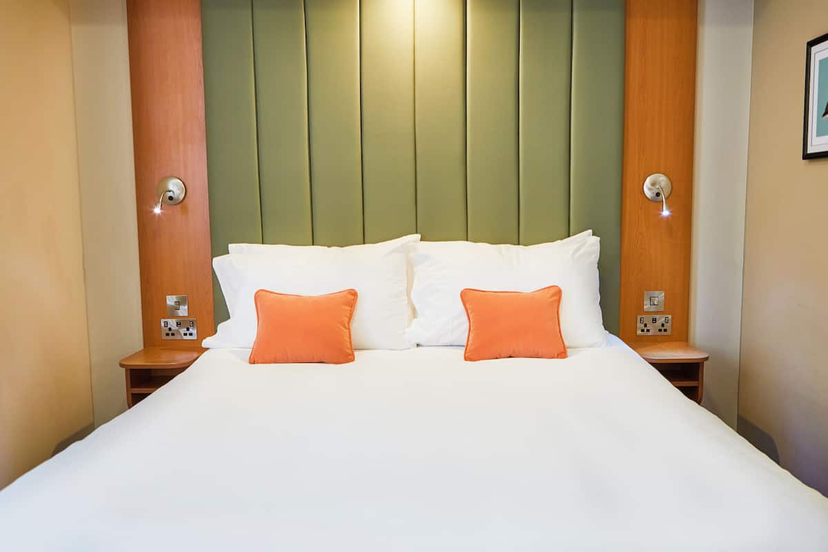 Une chambre d’hôtel avec un grand lit, deux oreillers orange, une tête de lit en bois et des tables de chevet avec des lampes.