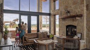A spacious living area with a stone fireplace, large windows, and a view of giraffes outside. - Longneck Manor (Fredericksburg)