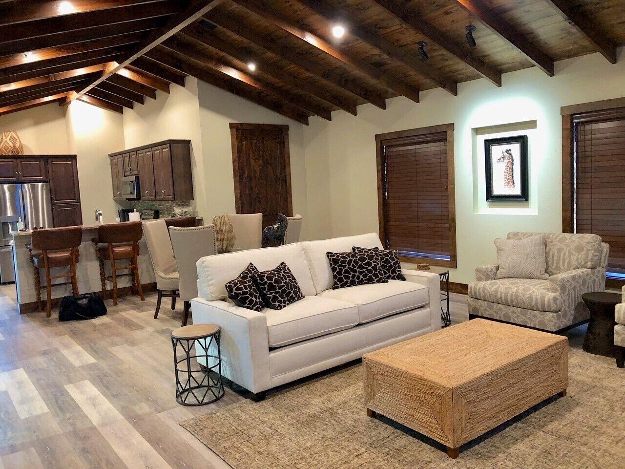 A modern living room with a white sofa, two patterned armchairs, a wooden coffee table, and a kitchen area with dark wooden cabinets and a stainless steel refrigerator.