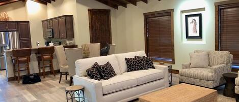 A modern living room with a white sofa, two patterned armchairs, a wooden coffee table, and a kitchen area with dark wooden cabinets and a stainless steel refrigerator.