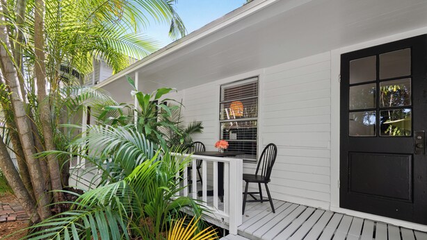 A white wooden porch with a black door, a small table with chairs, and lush tropical plants. - Charming 1-bed cottage in a Tropical Setting in West Palm Beach. Great location! (West Palm Beach)