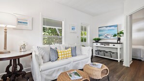 A modern living room with a white sofa, two wicker side tables, a white console table with baskets, and a painting of a boat. - Charming 1-bed cottage in a Tropical Setting in West Palm Beach. Great location! (West Palm Beach)