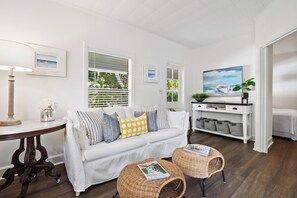 Un salon moderne avec un canapé blanc, deux petites tables d’appoint en osier, une console blanche agrémentée de paniers et un tableau représentant un bateau.